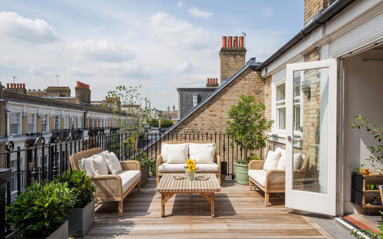 A beautiful and sunny rooftop terrace in London, furnished with stylish wicker outdoor seating and potted plants. This inviting roof garden features a wooden deck and offers a charming view over classic brick townhouses, providing a perfect urban oasis on a bright day.