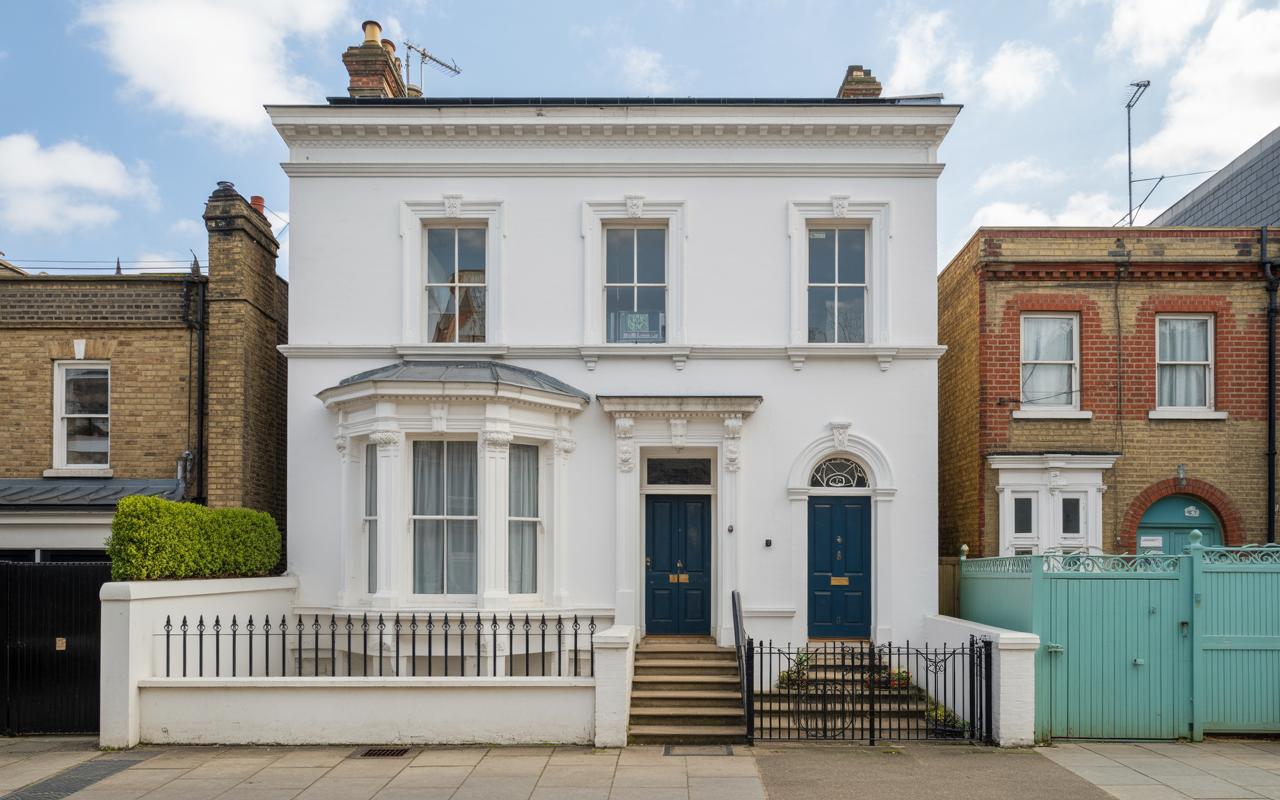 The stunning exterior of a large, white-painted Victorian detached house in London, featuring elegant architecture, a bay window, and two striking blue front doors. This property showcases classic British curb appeal with its pristine facade and iron railings.