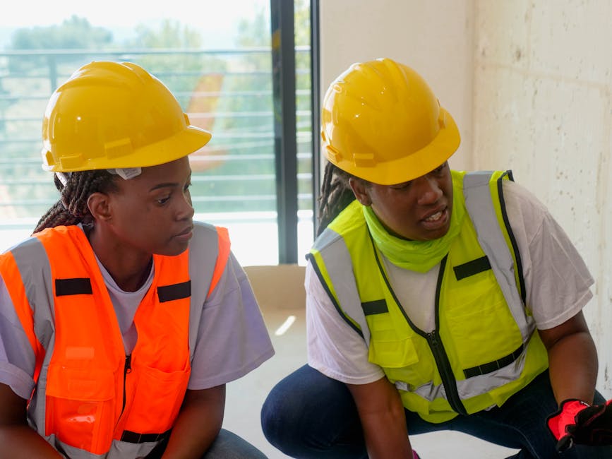 Two engineers wearing protective gear and hardhats discuss a project inside a building under construction.