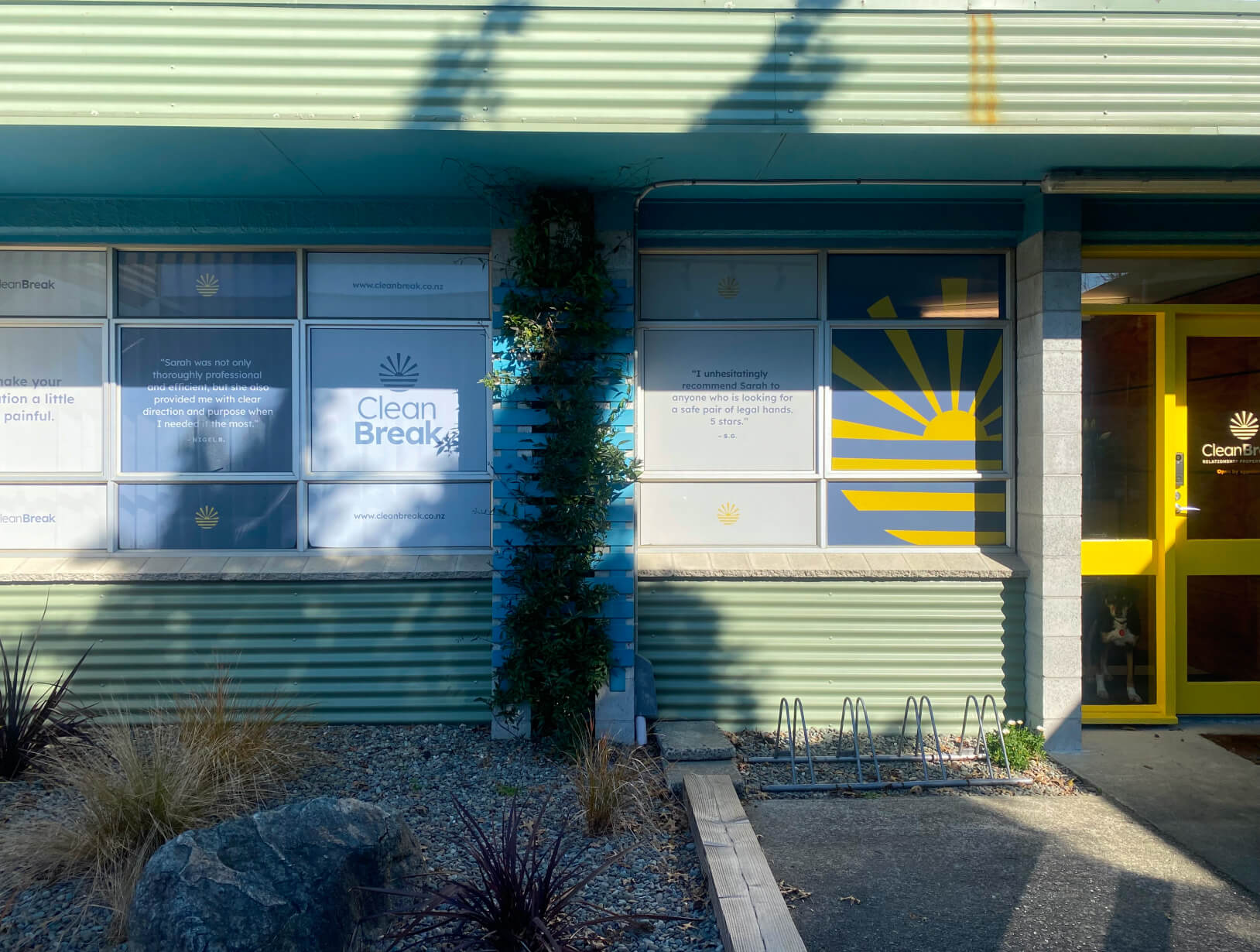 A building exterior with green corrugated siding, large windows displaying Clean Break signage, and a yellow sunburst design on the door and window.