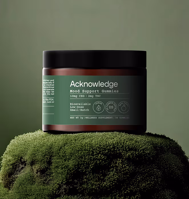 A dark amber jar of Acknowledge Mood Support Gummies with a green label sits on moss. The label highlights CBD and THC content, vegan, and small-batch qualities.