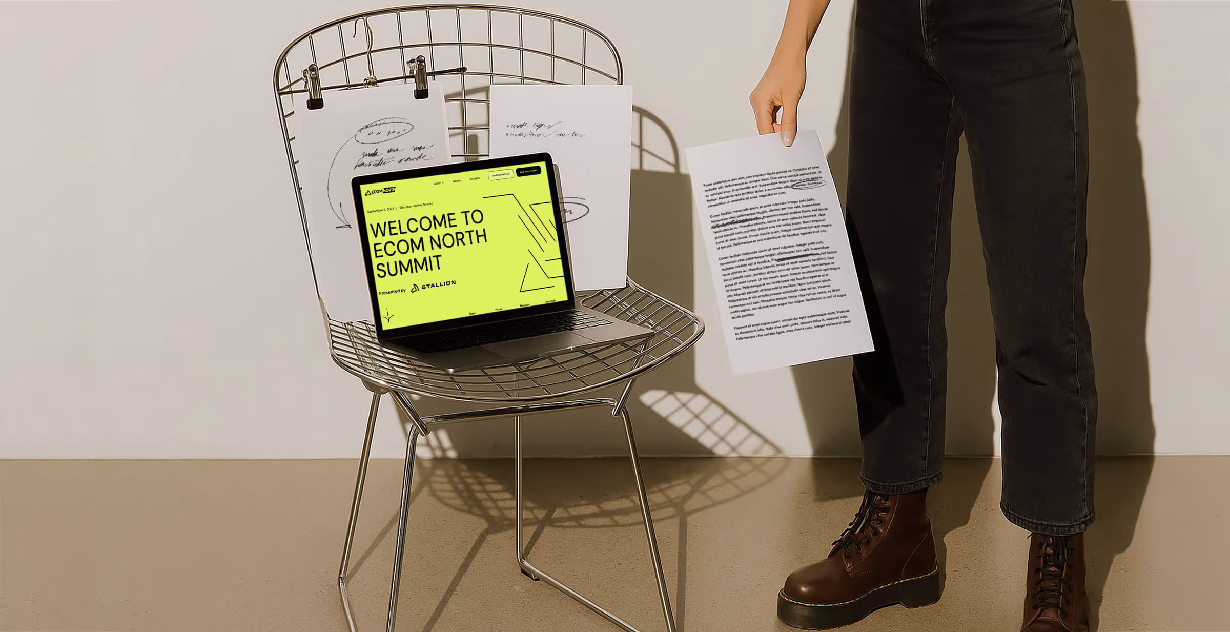 A laptop on a wire chair displays a bright yellow screen reading "WELCOME TO ECOM NORTH SUMMIT," with papers clipped above and one held by a person standing nearby.