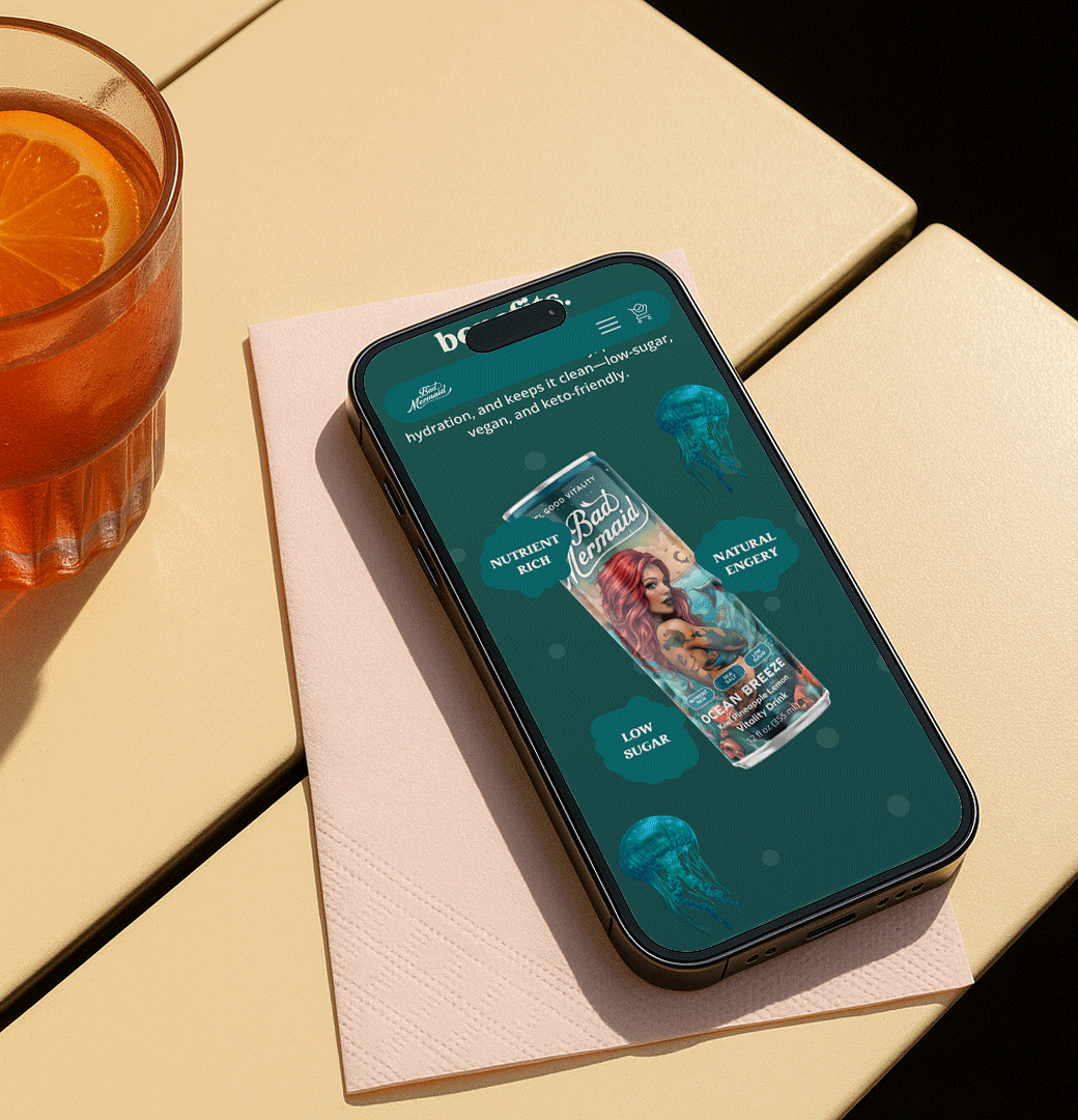 Smartphone on a napkin displaying a Bad Mermaid beverage can design with wellness highlights, next to a glass of orange drink.