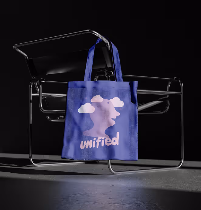 A blue tote bag with a pink abstract face-and-clouds illustration and the word "unified" printed on it, hanging from a modern chrome chair against a dark background.
