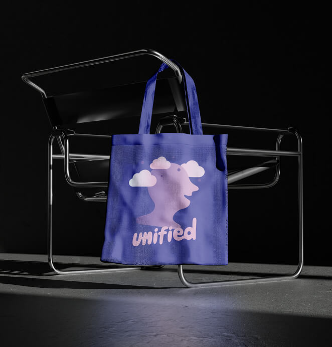 A blue tote bag with a pink abstract face-and-clouds illustration and the word "unified" printed on it, hanging from a modern chrome chair against a dark background.