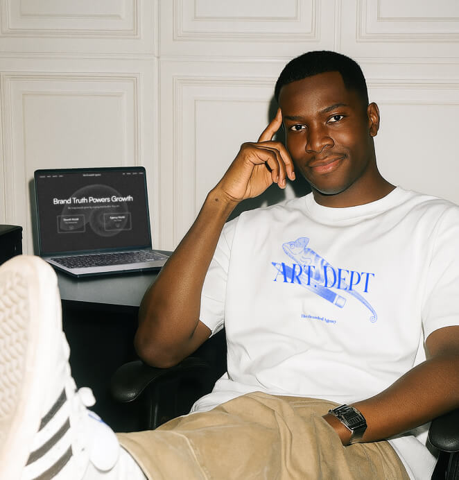 A man in a white t-shirt with a blue “Art Dept” design sits relaxed with one foot up, smiling at the camera. A laptop behind him displays the text “Brand Truth Powers Growth.”