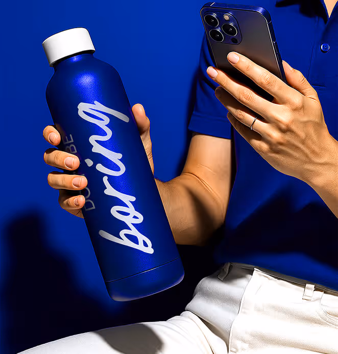 A close-up of hands holding a blue metallic water bottle with the word “boring” printed in large script and a smartphone, against a bold blue background.