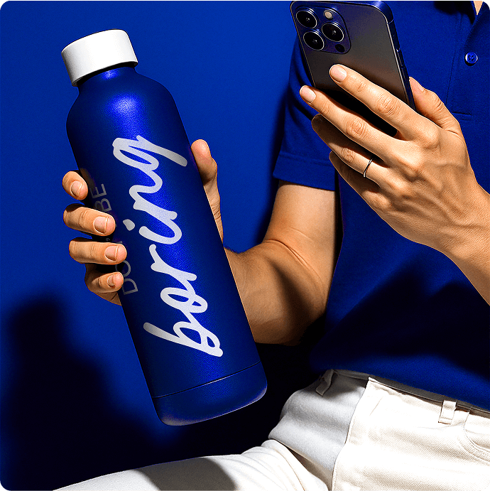 A close-up of hands holding a blue metallic water bottle with the word “boring” printed in large script and a smartphone, against a bold blue background.