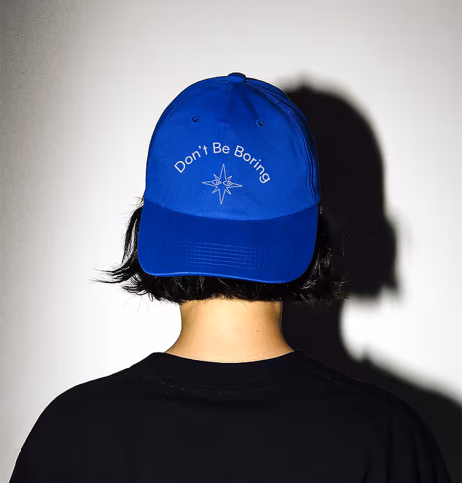Rear view of a person wearing a blue cap embroidered with the phrase “Don’t Be Boring” above a compass star graphic, set against a shadowed wall.