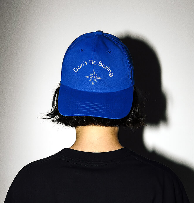Rear view of a person wearing a blue cap embroidered with the phrase “Don’t Be Boring” above a compass star graphic, set against a shadowed wall.