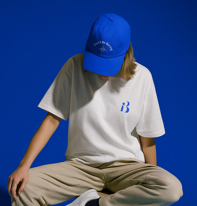 A person sits cross-legged wearing a white t-shirt with a blue “B” logo and a blue cap embroidered with “Don’t Be Boring,” against a matching deep blue backdrop.