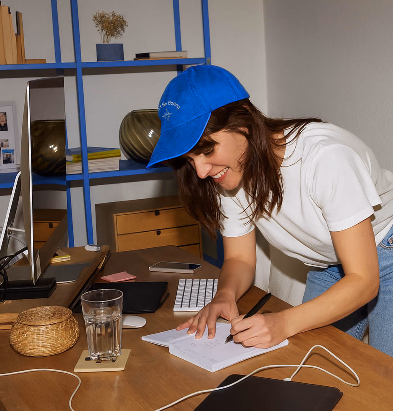 Person writing at a desk – A woman in a white t-shirt and blue cap that reads “Don’t Be Boring” leans over a desk, writing in a notebook. A desktop computer, keyboard, and glass of water are visible on the desk.