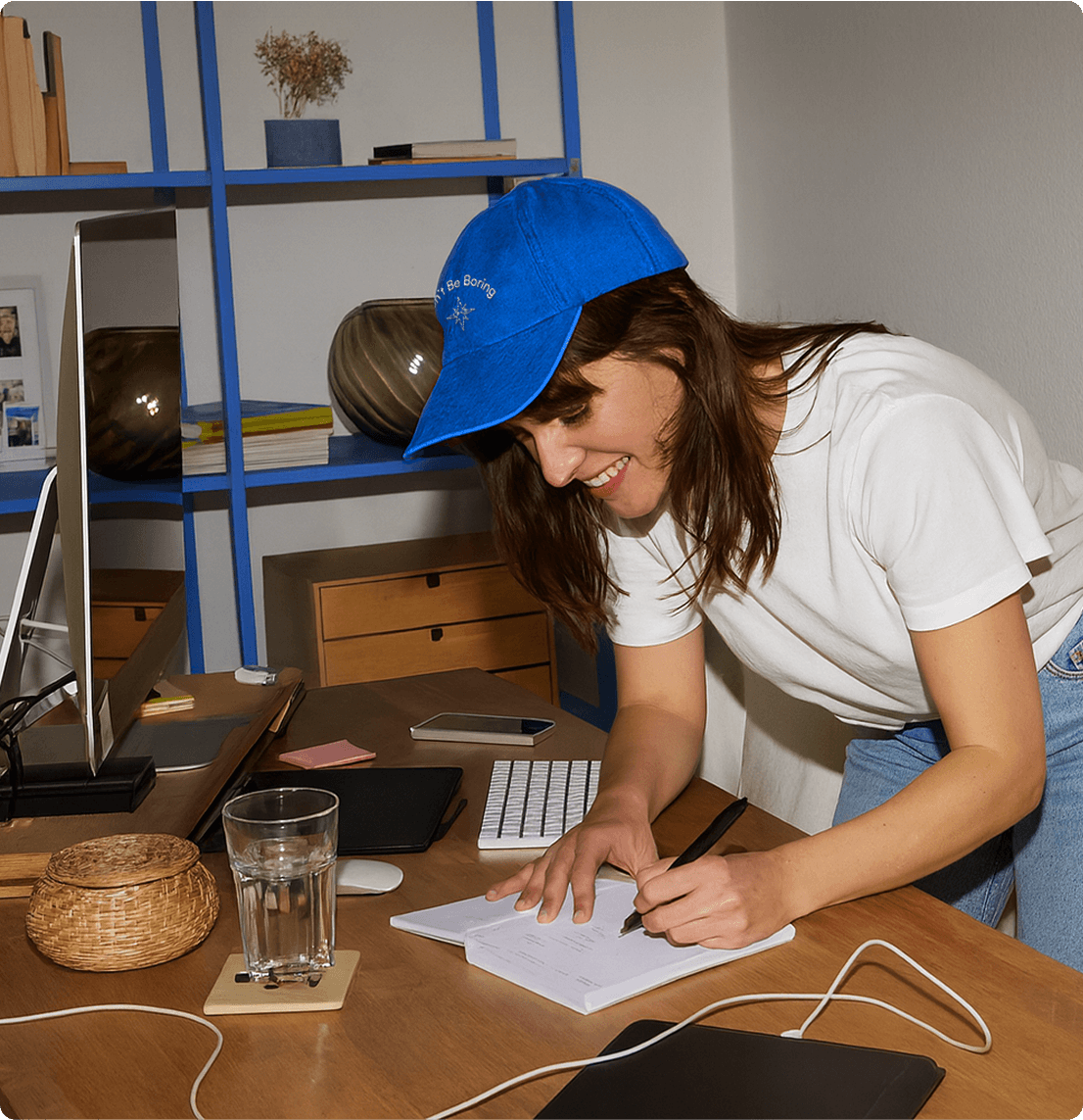 Person writing at a desk – A woman in a white t-shirt and blue cap that reads “Don’t Be Boring” leans over a desk, writing in a notebook. A desktop computer, keyboard, and glass of water are visible on the desk.