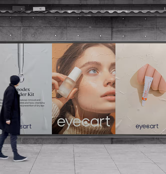 A person walks past a large outdoor billboard featuring Eyecart ads. The posters show skincare products, close-up imagery, and soft neutral tones.