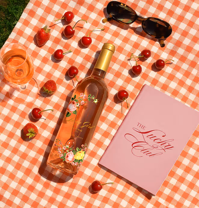 A picnic setup on a red-and-white checkered cloth with a bottle of Lucky Girl Rosé wine, fresh cherries, strawberries, sunglasses, and a pink booklet titled “The Lucky Club.”
