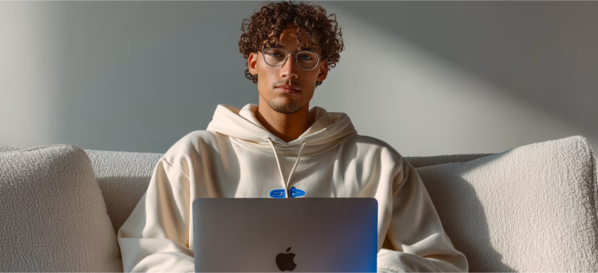 A young man in glasses and a cream hoodie sits on a sofa working on a MacBook, lit by natural sunlight against a neutral background.