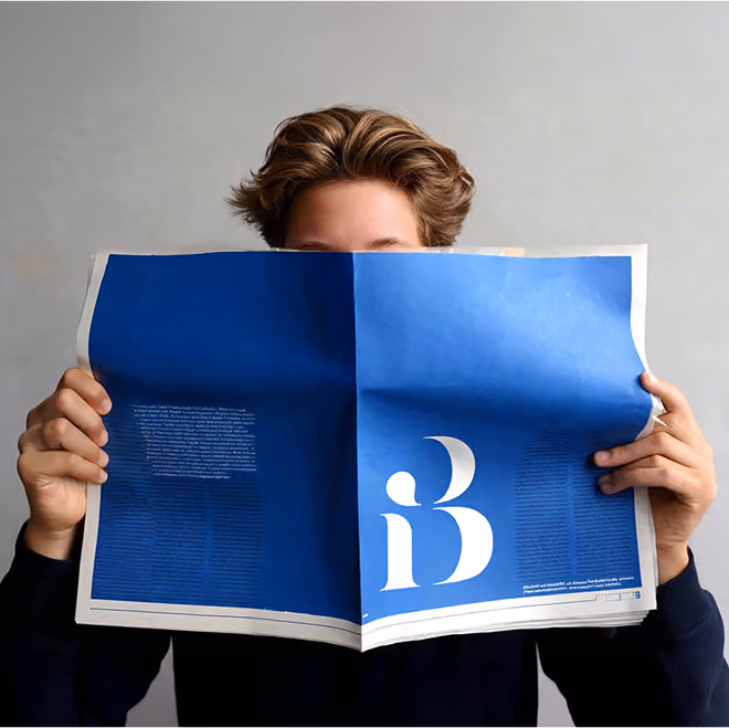 A person holding up a blue newspaper that covers their face, featuring a large white “B” logo on the front page.