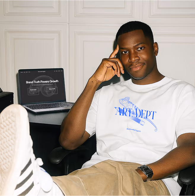 A man sitting confidently at his desk with one hand on his temple, wearing a white “ART DEPT” t-shirt, with a laptop in the background displaying “Brand Truth Powers Growth.”