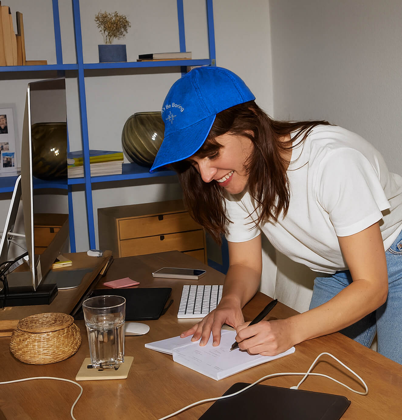 A woman wearing a bright blue cap and white t-shirt leans over a wooden desk, smiling as she writes in a notebook surrounded by work essentials like a glass of water, keyboard, and phone.