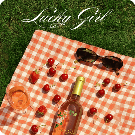 Picnic scene with rosé wine bottle, cherries, sunglasses, and gingham blanket on grass, styled lifestyle branding image for Lucky Girl.