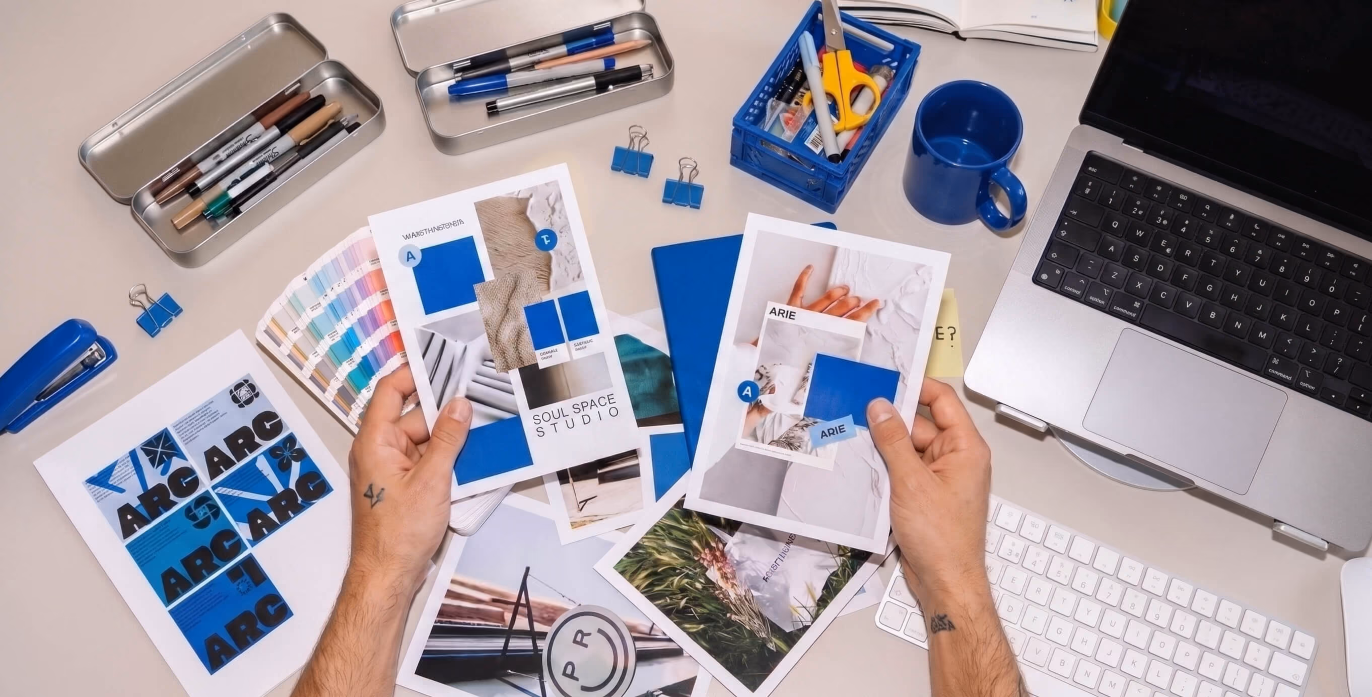 Overhead view of a designer reviewing brand concepts and visual identity materials at a creative workspace with color swatches, stationery, and a laptop.