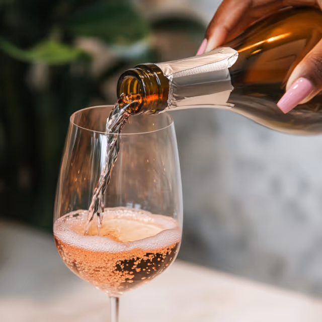 A close-up of sparkling rosé wine being poured into a glass, with the bubbles forming at the surface, held by a hand with manicured nails.