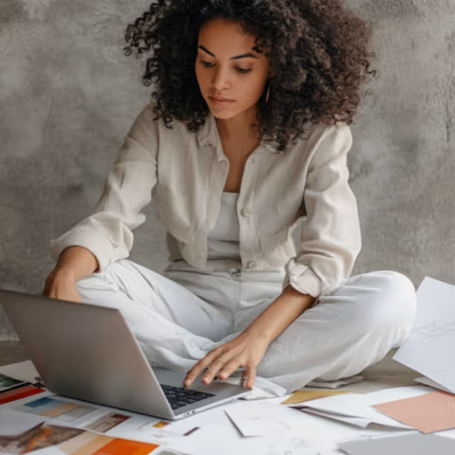 A woman working on her laptop, surrounded by papers and design materials. She is seated on the floor, deep in concentration, reflecting creativity and remote work flexibility.