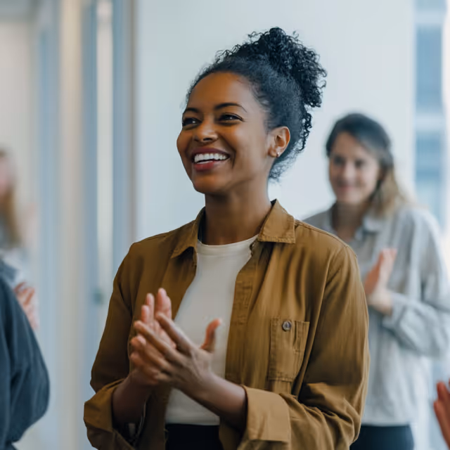 Smiling woman in a brown jacket clapping in a modern office environment, surrounded by colleagues in the background.