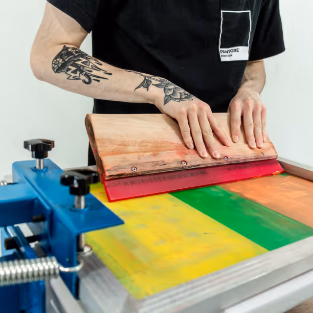 Close-up of a tattooed individual pressing ink through a screen printing frame, producing multicolored prints.