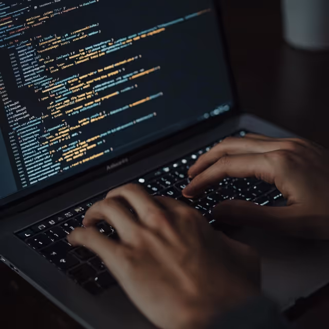 Close-up of hands typing code on a laptop keyboard in a dimly lit environment.