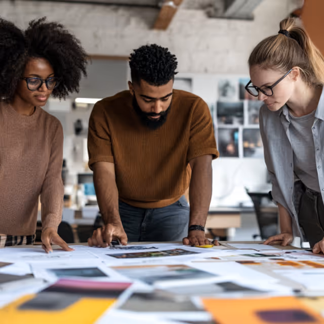 Three creative professionals analyzing printed design concepts spread across a large table in a collaborative studio setting.