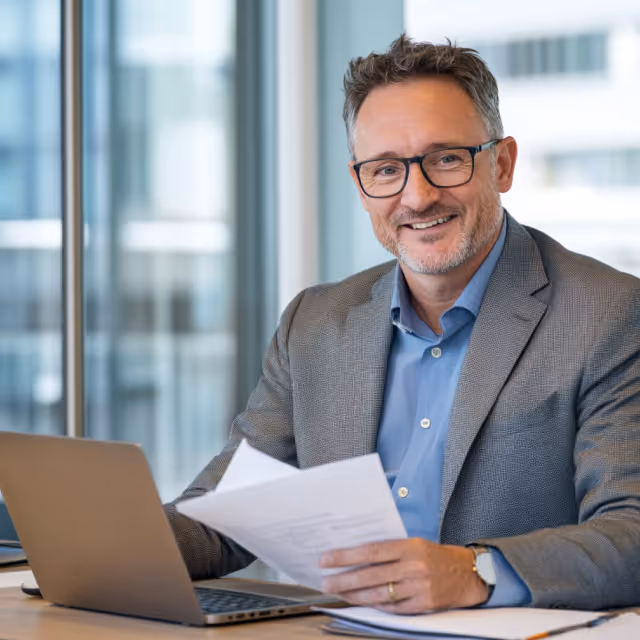 A smiling man in glasses and a blazer sits at his desk with a laptop and documents, looking directly at the camera in a bright office.