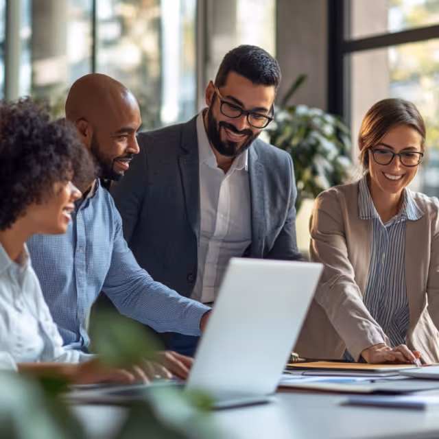 Four diverse colleagues gather around a laptop, smiling and discussing ideas in a modern office with natural light.
