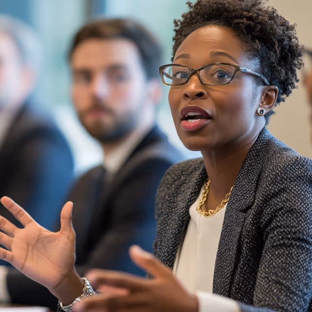 A focused woman in glasses speaks during a business meeting, gesturing with her hands, with blurred colleagues seated behind her.