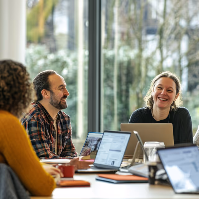 A diverse group of coworkers smiling and laughing around a table with laptops, highlighting collaboration and positive workplace culture.