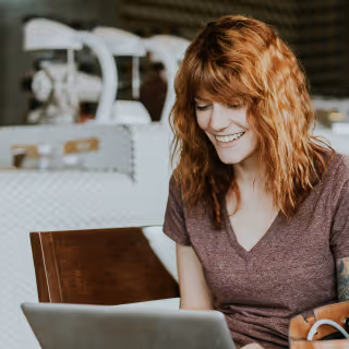 A young woman with red hair smiles while working on a laptop in a cozy café environment, symbolizing casual and creative work culture.