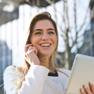 A cheerful young woman in a white blazer smiles while holding a tablet and talking on a phone, standing outdoors in a modern urban setting.