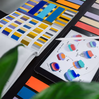 A close-up of a tablet and an open book showing colorful design swatches and 3D shape sketches, representing a creative design process.