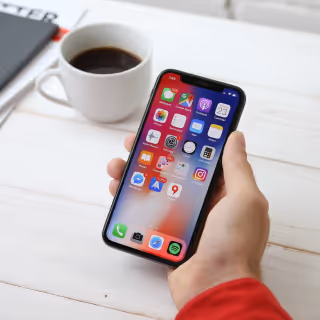 A hand holding a smartphone displaying the home screen filled with colorful app icons, placed beside a cup of coffee on a white wooden table.