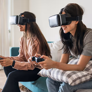 Two women wearing VR headsets sit on a couch, smiling and playing a video game with controllers in hand, immersed in virtual reality.