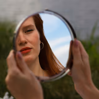 A person holds a round mirror revealing only part of a woman's face with red lipstick, symbolizing self-reflection and identity.