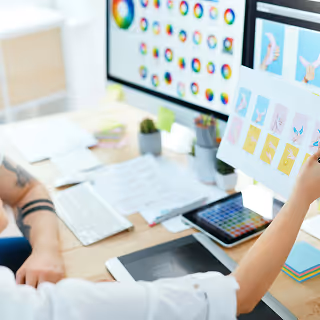 Creative professional examining printed brand icon concepts in front of dual monitors showing colorful digital palettes and design software.
