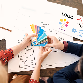 Close-up of two people reviewing a colorful swatch fan and brand design sketches, including logo and website wireframes on a wooden table.