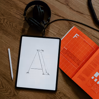 Top-down view of a tablet showing a typographic letter “A” on grid paper, with a stylus, open typography book, and headphones beside it.