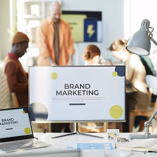 Office desk with a computer and laptop screen displaying “Brand Marketing,” with blurred coworkers collaborating in the background.
