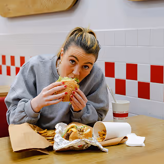 Woman sitting at a fast food restaurant enthusiastically biting into a large hamburger, with fries and a drink on the table.