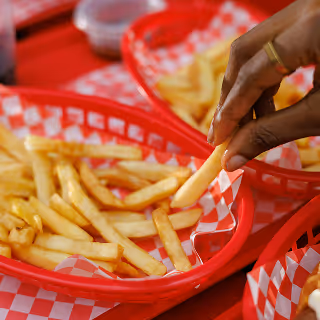Close-up of a hand picking up a French fry from a red plastic food basket lined with red-checkered paper.