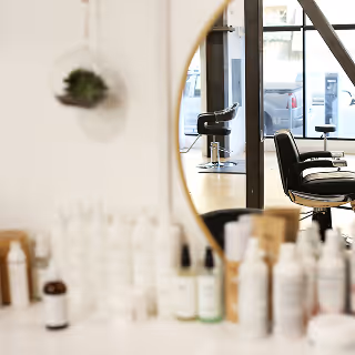 A salon countertop filled with white bottles of hair products, with a salon chair visible in a round mirror's reflection.