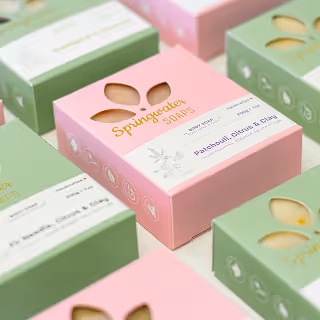 A close-up of pastel pink and green soap boxes with floral cutouts labeled "Springwater Soaps," arranged neatly for product display.