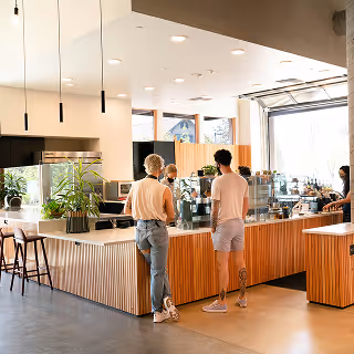 Modern coffee shop interior featuring wood-accented counters, indoor plants, and customers standing at the barista counter under soft lighting.